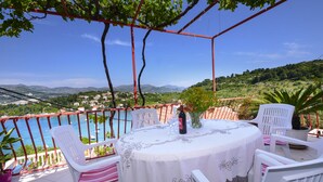 Een ronde tafel met een wit tafelkleed en stoelen met roze en witte kussens, op een balkon met uitzicht op een wateroppervlak en een bergachtig landschap.