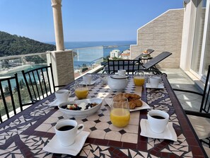Ett frukostbord med croissanter, frukt, kaffe och apelsinjuice på en balkong med utsikt över kusten.