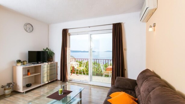 A living room with a glass coffee table, a brown sofa with an orange pillow, a TV on a wooden cabinet, and a balcony with a view of the sea. - Guest House Fontana - Two Bedroom Apartment with Balcony and Sea View (Mlini)