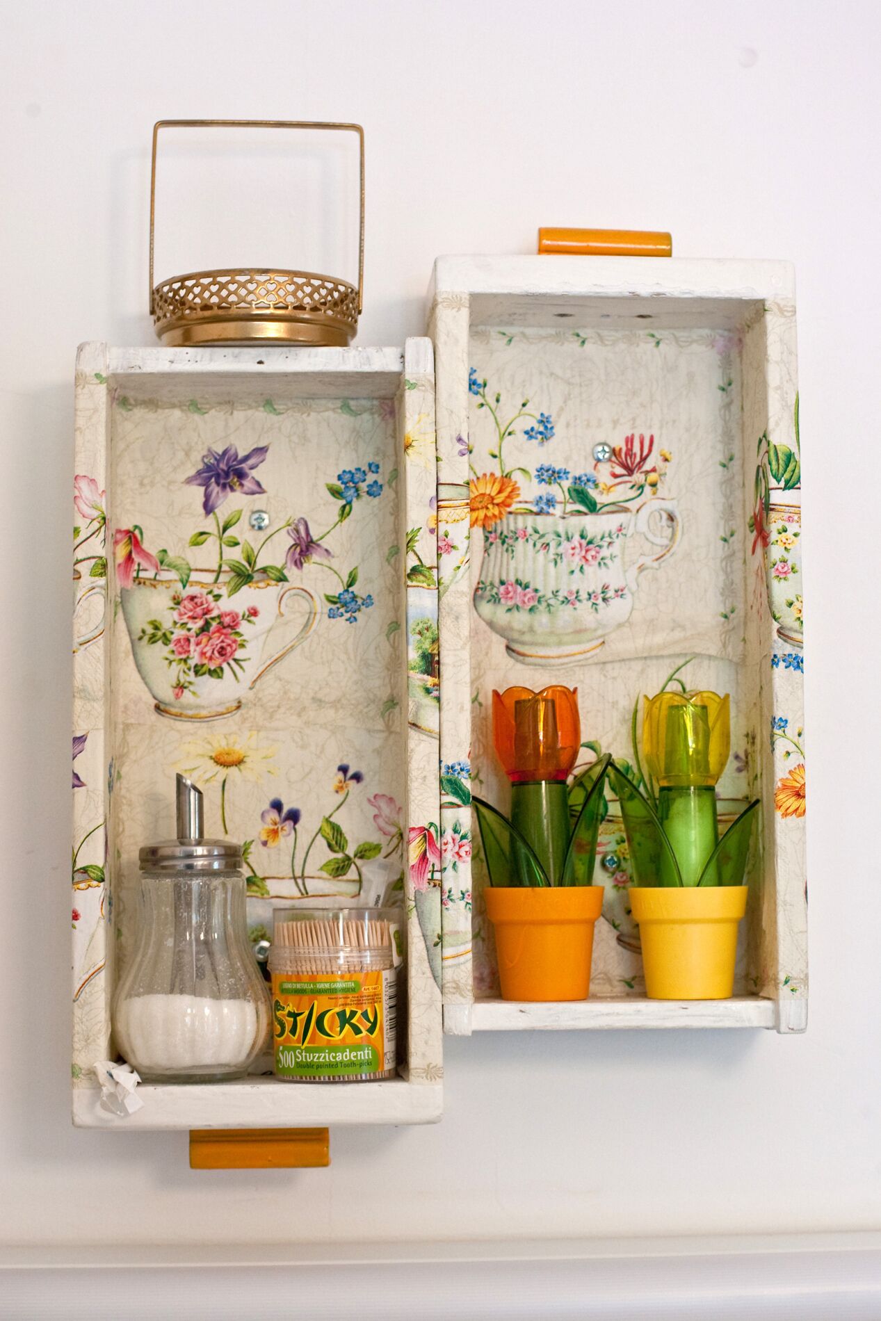 Dos cajas decorativas abiertas con motivos florales y asas, que contienen un frasco de vidrio, un soporte para cepillos de dientes y dos macetas.