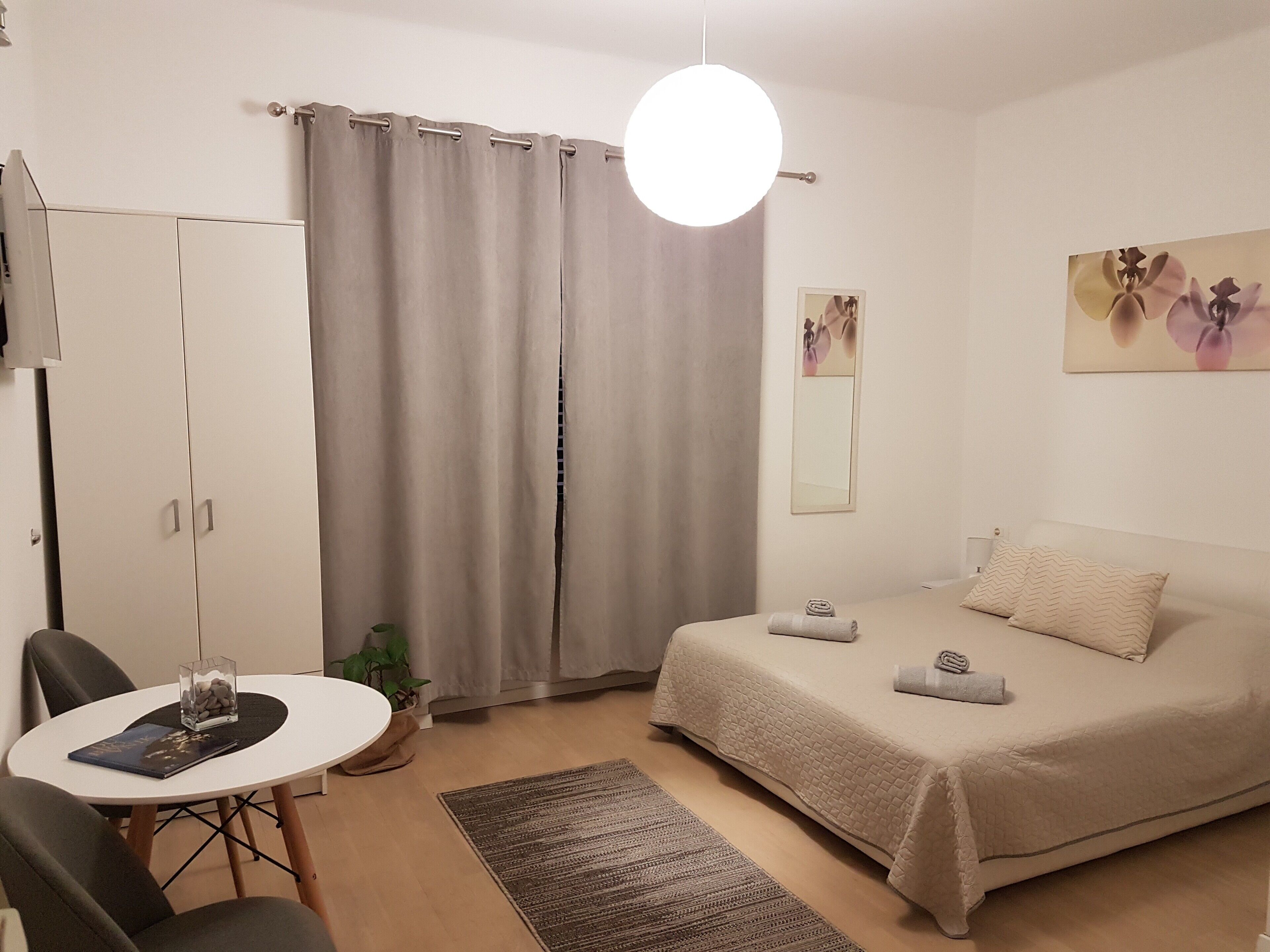 A bedroom with a bed, a desk with a chair, a wardrobe, a potted plant, and a painting on the wall.