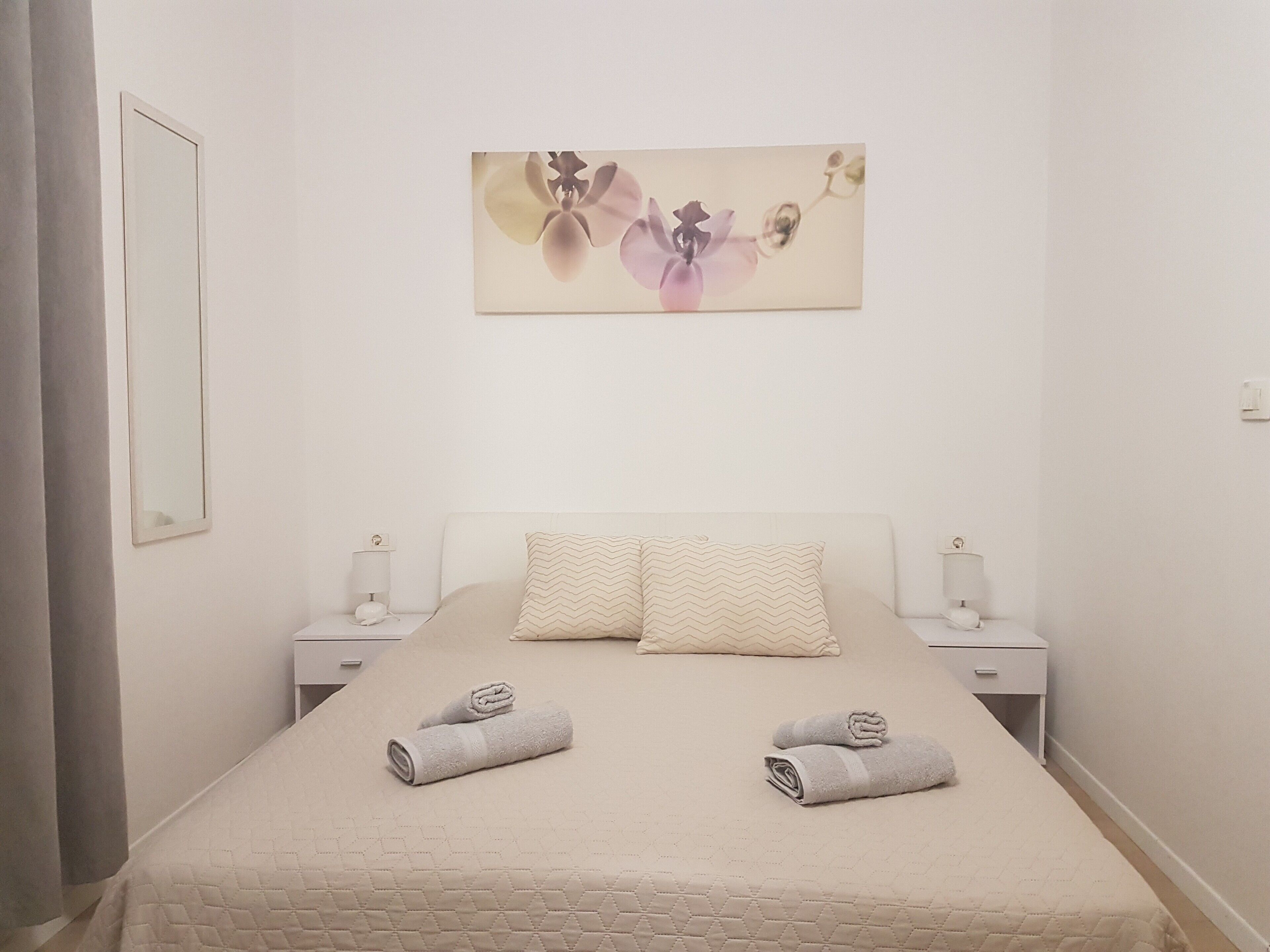 A bedroom with a bed, two nightstands, a wall-mounted mirror, and a floral artwork.