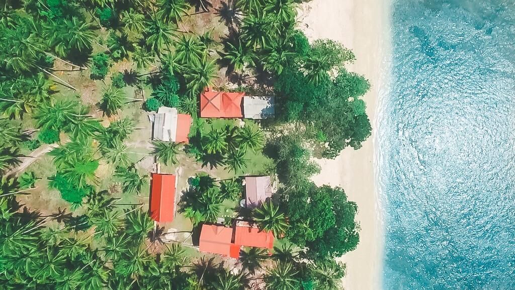 An aerial view of a coastal area with several red-roofed structures surrounded by palm trees, adjacent to a sandy beach and turquoise ocean.