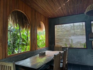 A wooden dining area with a bench and chairs, large windows with blinds, and a view of greenery outside. - Krui Surfing (South Krui)