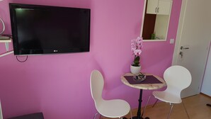 A room with a pink wall, a flat-screen TV, a small table with chairs, a potted orchid, and a mirror. - Guest House Avdic - Studio Apartment with Balcony (Dubrovnik)