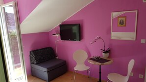 A room with a pink wall, a grey sofa, a TV, a small table with a potted plant, and two white chairs. - Guest House Avdic - Studio Apartment with Balcony (Dubrovnik)