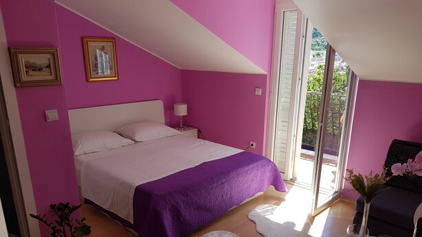 A bedroom with a bed, a nightstand, a lamp, a potted plant, and a window with a view of greenery. - Guest House Avdic - Studio Apartment with Balcony (Dubrovnik)