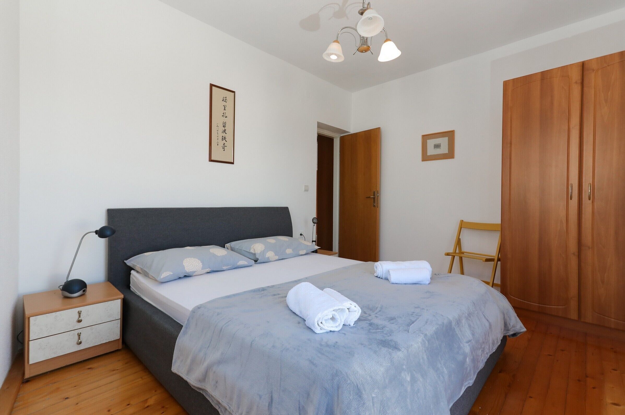 Un lit bien fait, avec une couverture grise et des oreillers, une table de chevet avec une lampe, une armoire en bois et un tableau encadré au mur.