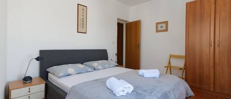 Un lit bien fait, avec une couverture grise et des oreillers, une table de chevet avec une lampe, une armoire en bois et un tableau encadré au mur.