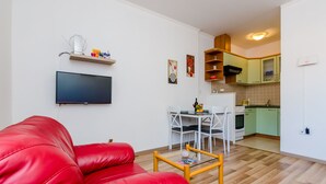 Un espace de vie compact comprenant un canapé en cuir rouge, un téléviseur à écran plat, une table à manger avec des chaises et une kitchenette dotée de meubles de couleur vert clair.