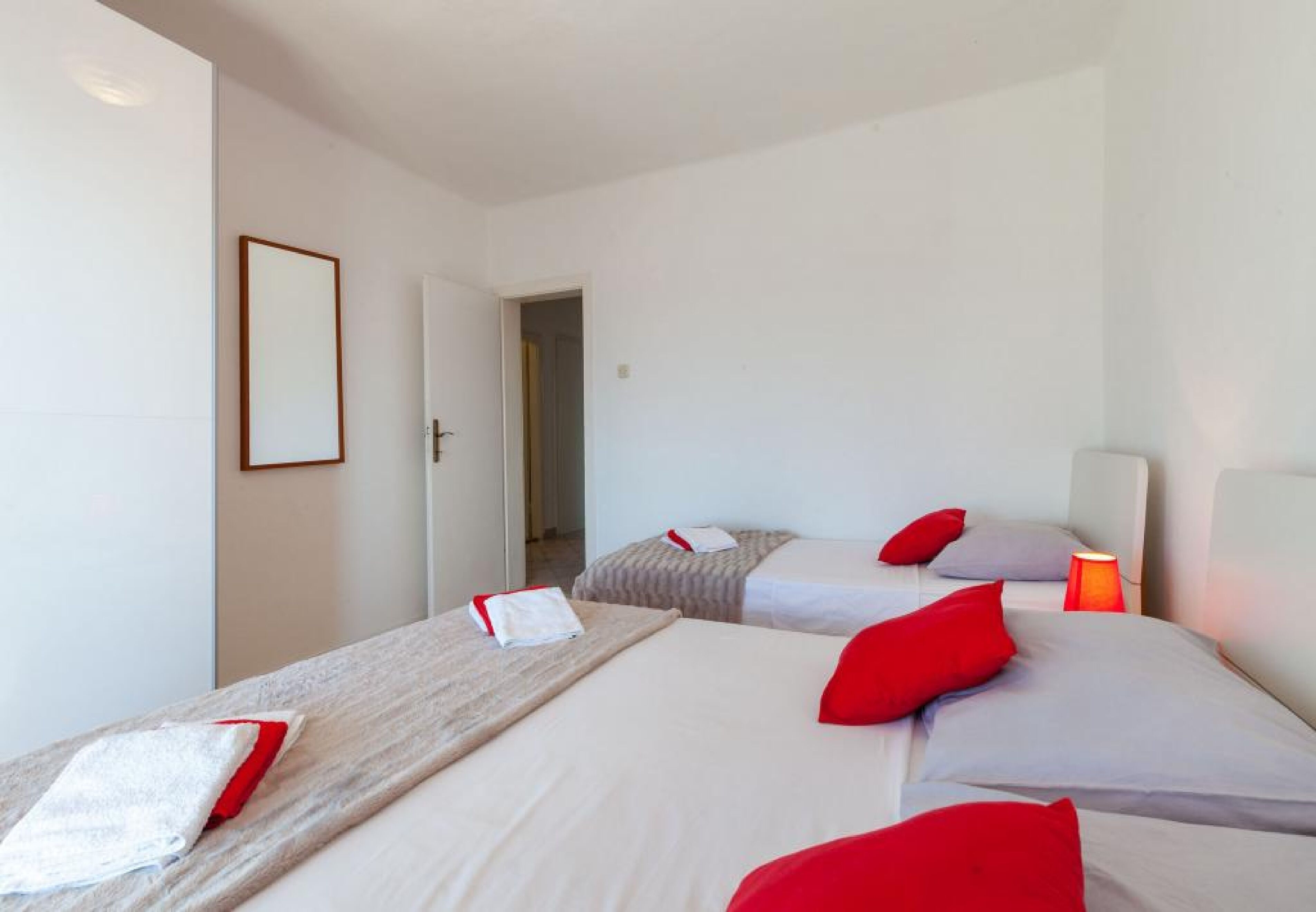 A hotel room with two beds, each with red and white bedding, a bedside table with a lamp, and a door leading to another room.