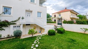 Ein zweistöckiges Gebäude mit weißer Fassade, einem Balkon und einem kleinen Garten mit einem Kiesweg.