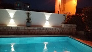 Ein Schwimmbecken mit Fliesenrand, eine Ziegelwand mit Topfpflanzen und ein Gebäude mit beleuchteten Fenstern im Hintergrund.