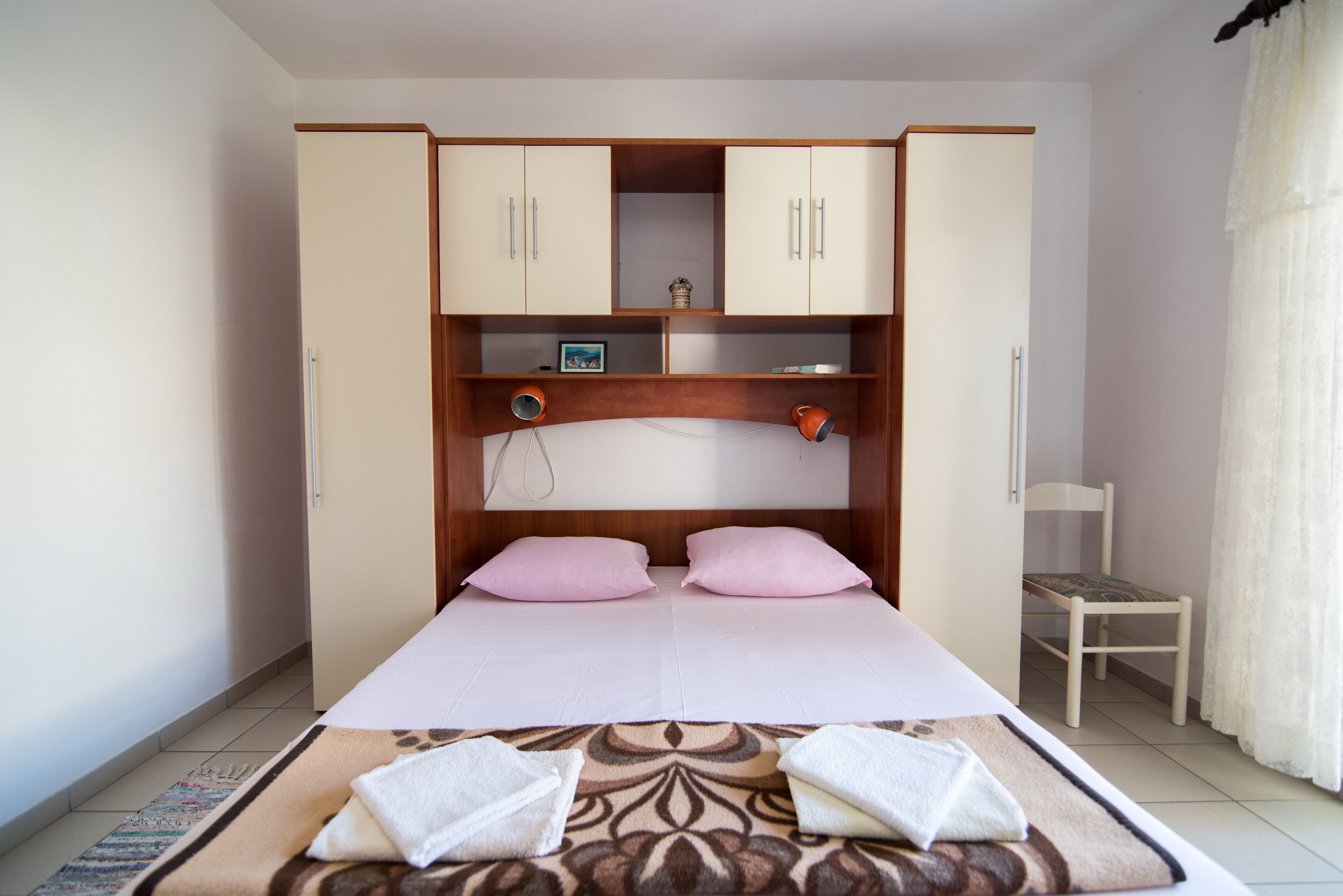 Ein Schlafzimmer mit einem Bett, zwei rosafarbenen Kissen, einem Holzschrank, einem Stuhl und einem kleinen Tisch mit einem Bilderrahmen.