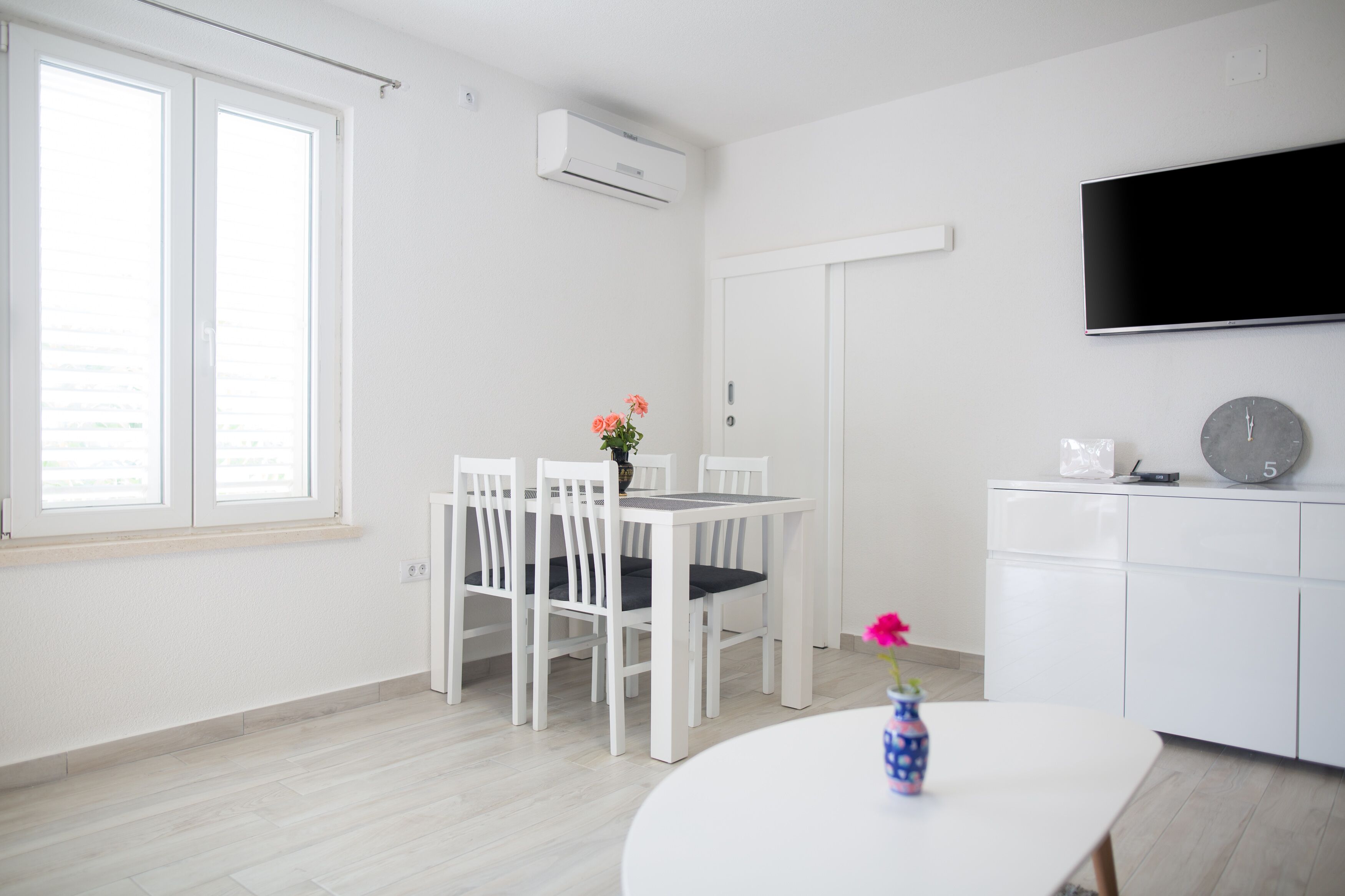 A modern, minimalist room with a dining table and chairs, a flat-screen TV, and a clock on the wall.