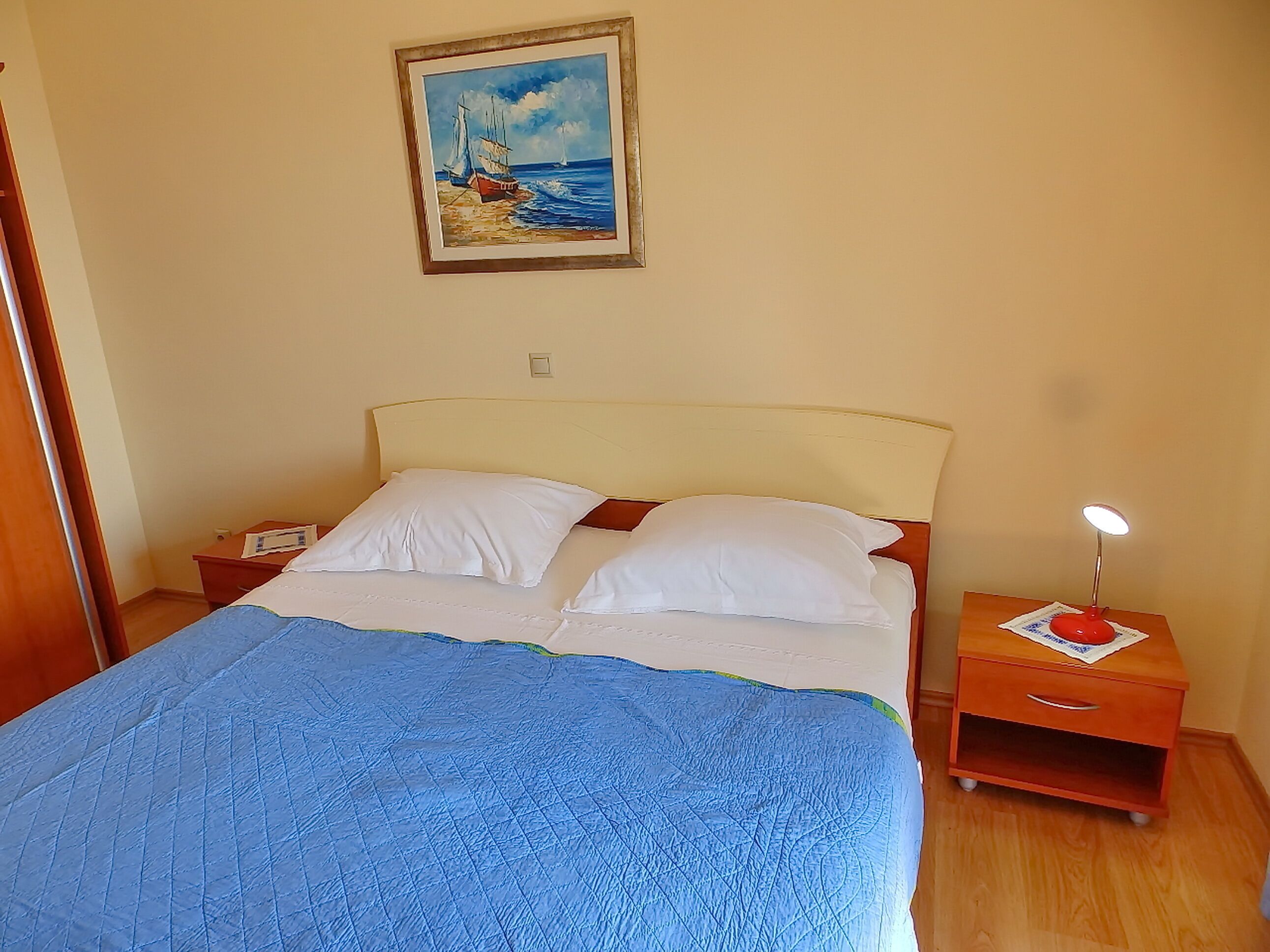 A bedroom with a bed, two pillows, a nightstand with a lamp, a painting on the wall, and a wooden floor.