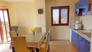 Un coin repas avec une table, des chaises et un vase de fleurs. Il y a une fenêtre avec des stores, un four à micro-ondes et des placards dans la cuisine.