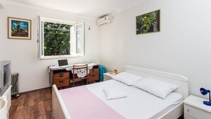 Ein Schlafzimmer mit Bett, Schreibtisch mit Laptop, Stuhl, Heizkörper und einem Fenster mit Blick ins Grüne.