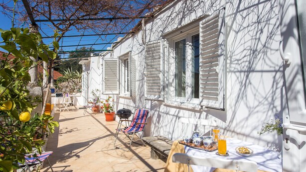 Eine Terrasse mit Esstisch, Sitzgelegenheiten im Freien und Blick auf ein weißes Gebäude mit Fensterläden.