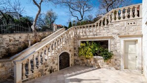 Un escalier en pierre mène à une entrée fermée par un portail, l’ensemble étant entouré de murs de pierre et d’arbres.