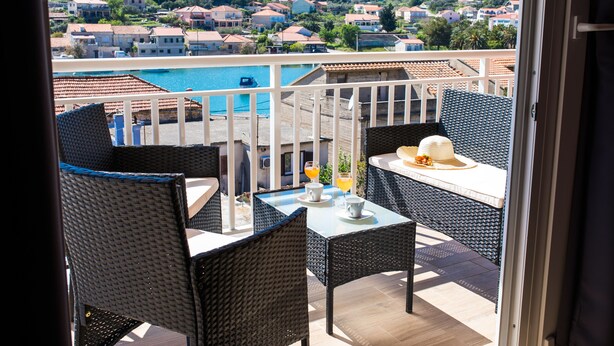 Ein Balkon mit Korbmöbeln, einem Tisch und Blick auf ein Schwimmbad und Häuser am Hang.