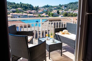 A balcony with wicker furniture, a table, and a view of a swimming pool and hillside houses.