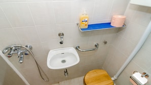 Ein kleines Badezimmer mit Waschbecken, Toilette und Dusche. Ein Ablageregál mit einer Seifenflasche und einer Packung Toilettenpapier.