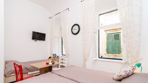 Una habitación con dos camas, un televisor montado en la pared, una mesita con una silla, un ventanal con contraventanas verdes y un reloj.