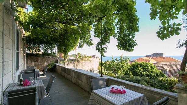 An outdoor patio with tables and chairs, a stone wall, and a view of the sea and buildings in the background. - Villa Iveta - Twin Room (Ground Floor) - Martina (Dubrovnik)