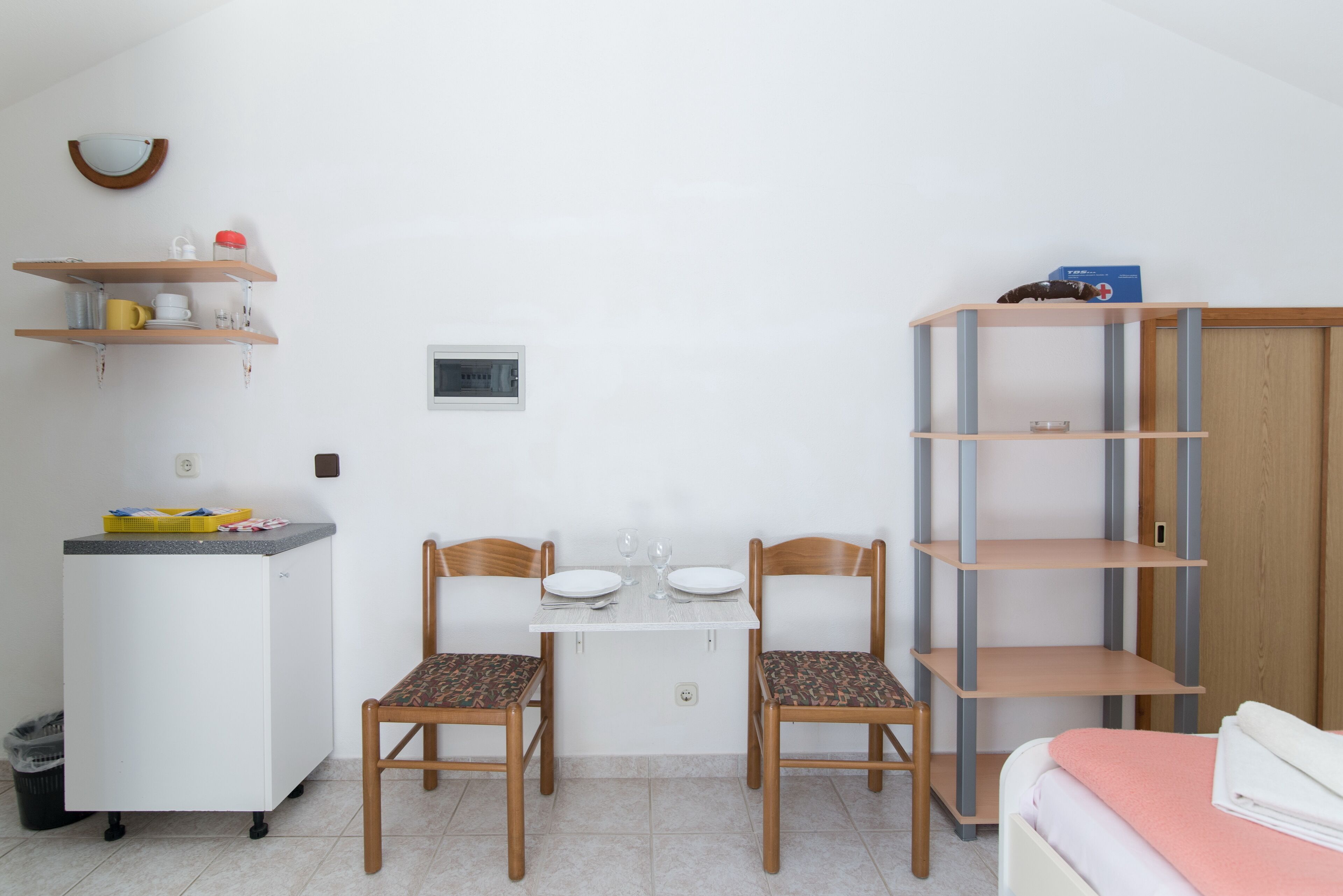 A small, clean room with a kitchenette, two wooden chairs, a white table, and a bed with a pink blanket.