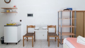 Ein kleiner, sauberer Raum mit einer Küchenzeile, zwei hölzernen Stühlen, einem weißen Tisch und einem Bett mit rosa Decke.