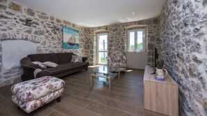 Una sala de estar con paredes de piedra, un sofá marrón, una mesa de centro de vidrio y un reposapiés con estampado floral.