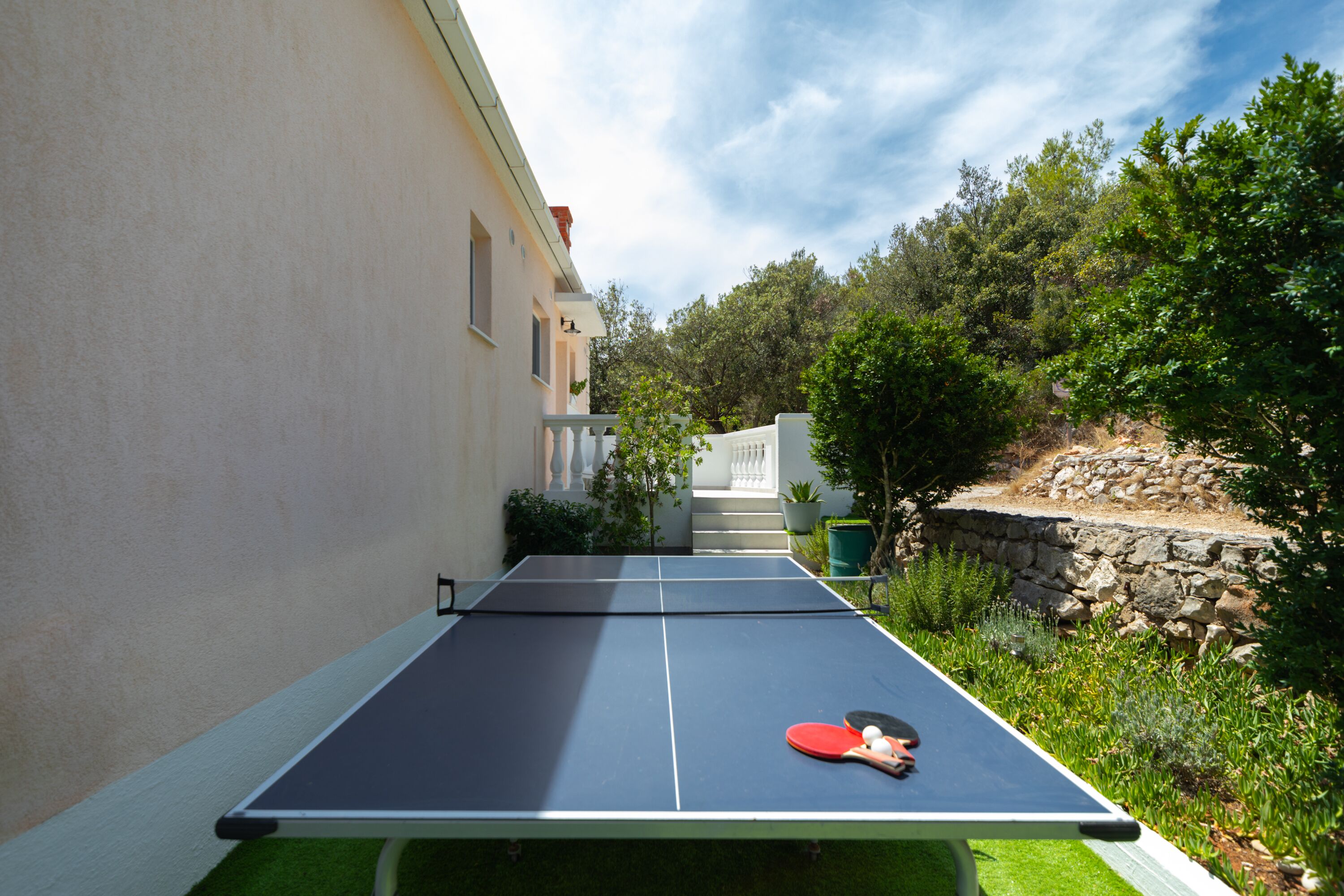 A ping pong table in an outdoor setting with a stone wall and greenery.