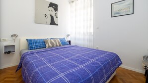 A bedroom with a bed, a nightstand, a wall-mounted lamp, a window with curtains, and a framed picture on the wall. - Old City Center Suite - One Bedroom Suite (Split)