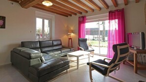 A living room with a black leather sofa, a wooden coffee table, two chairs, a TV, and a window with pink curtains.