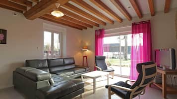 Un salon avec un canapé en cuir noir, une table basse en bois, deux fauteuils, un téléviseur et une fenêtre ornée de rideaux roses.