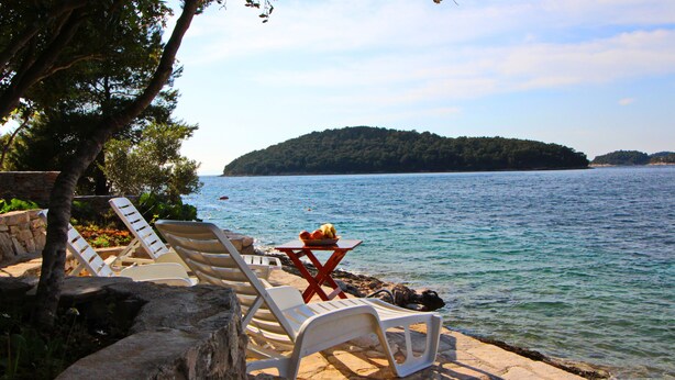 A stone deck with white lounge chairs and a table with fruit, overlooking a calm lake with a forested island in the distance. - Holiday Home Katica - One Bedroom Holiday Home with Terrace and Sea View (Vela Luka)