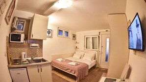 A compact hotel room with a kitchenette, a bed with a pink bedspread, a TV, and a small dining area. - Nika Family Apartments - Basic Studio Apartment with External Terrace and Sea View (A5) (Dubrovnik)