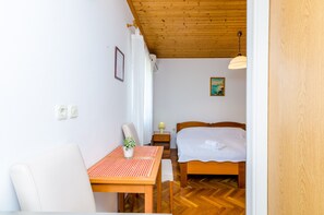 A room with a wooden ceiling, a bed with a white duvet, a small table with a plant, and a chair.