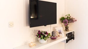 A flat-screen TV mounted on a white wall, a white shelf with a vase of flowers, a candle, and a framed photo.