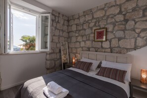 Ein Schlafzimmer mit Stehwand, einem großen Bett mit weißen und gestreiften Kissen, einem Nachttisch mit Lampe und einem Fenster mit Blick auf Palmen.