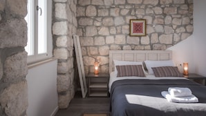 Quarto com parede de pedra, cama com almofadas, mesa de cabeceira com abajur e uma janela.