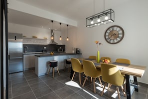 Een moderne keuken met een centraal kookeiland, roestvrijstalen apparatuur, hanglampen, en een eethoek met gele stoelen en een houten tafel.