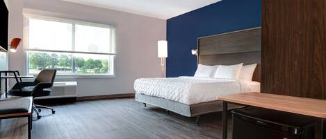 A hotel room with a bed, desk, chair, and a small refrigerator.