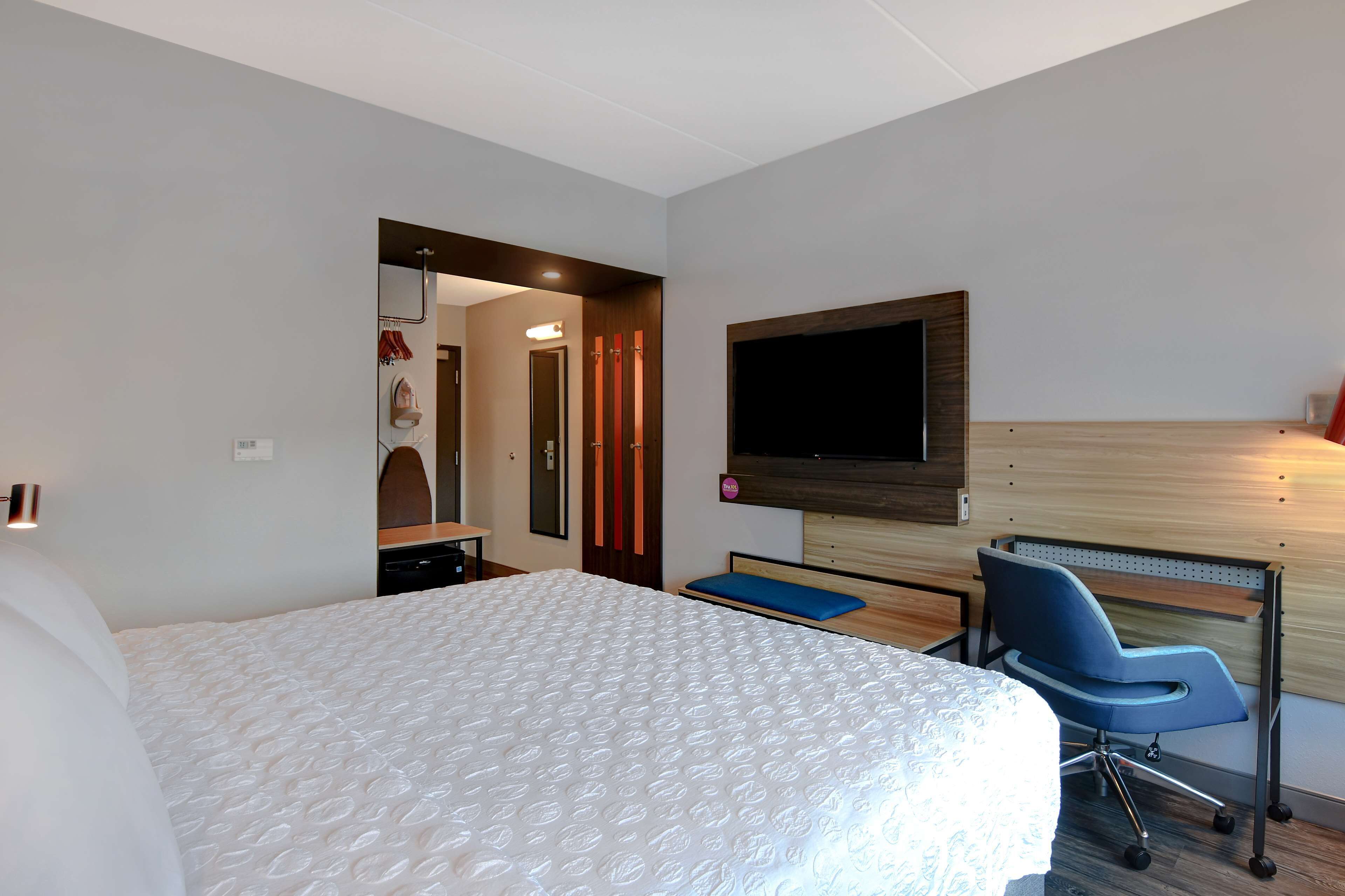 A hotel room with a large bed, a desk with a chair, a flat-screen TV mounted on the wall, and a wooden headboard.
