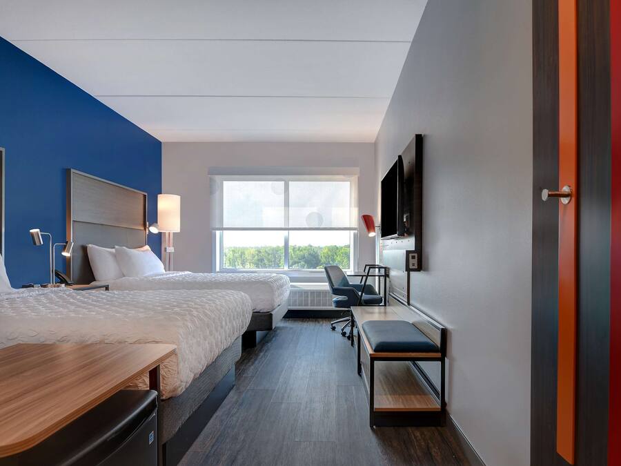 A modern hotel room with two beds, a desk, a chair, a TV, and a large window.