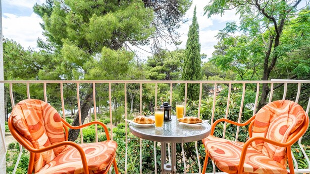 Un balcón con dos sillas naranjas, una mesita redonda con provisiones para el desayuno, y vistas a vegetación exuberante y árboles.