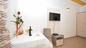 Ein Zimmer mit einem weißen Tisch, gedeckt mit einer Weinflasche, Gläsern und einer Rose. Ein Fernseher ist an der Wand montiert und ein Kleiderschrank steht in der Ecke.