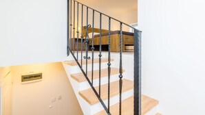 Una escalera con barandilla de metal negro y peldaños de madera que conduce a un dormitorio con cabecera de madera y una lámpara de noche.