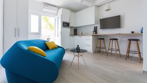 Una cocina moderna con armarios blancos, un microondas y un televisor de pantalla plana. Hay un sofá azul con cojines amarillos y una pequeña mesa redonda con taburetes.
